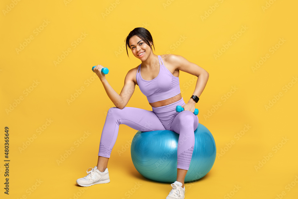 Happy black lady in sports clothes sitting on fitball and working out with dumbbells on yellow studio background Happy black lady in sports clothes sitting on fitball and working out with dumbbells on yellow studio background