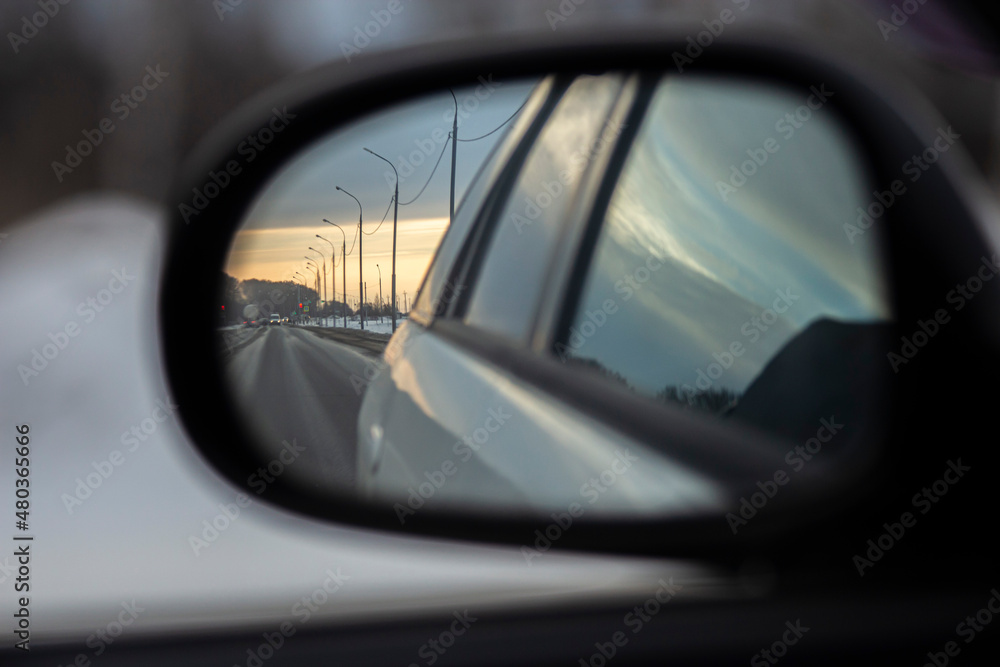 Reflection in the rearview mirror of a car. It displays a highway, lampposts along it, cars driving along the road, and in the background you can see the rays of the setting sun. 