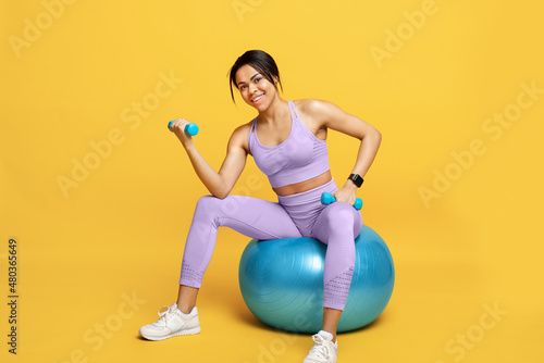 Happy black lady in sports clothes sitting on fitball and working out with dumbbells on yellow studio background