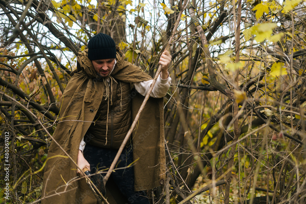 Portrait of serious tourist male wearing raincoat tent chopping small branches with small shovel in forest on overcast cold day. Concept of exploration, travel and adventure in nature. Portrait of serious tourist male wearing raincoat tent chopping small branches with small shovel in forest on overcast cold day. Concept of exploration, travel and adventure in nature.