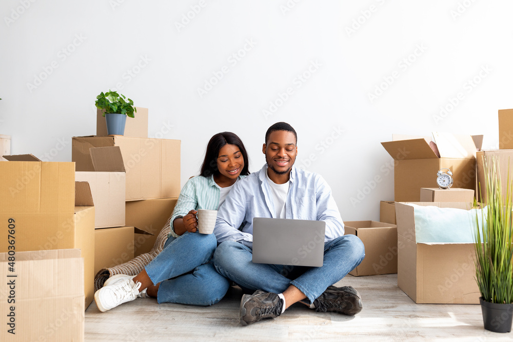 Happy african american spouses using laptop sitting among boxes after moving new home, browsing internet, free space Happy african american spouses using laptop sitting among boxes after moving new home, browsing internet, free space