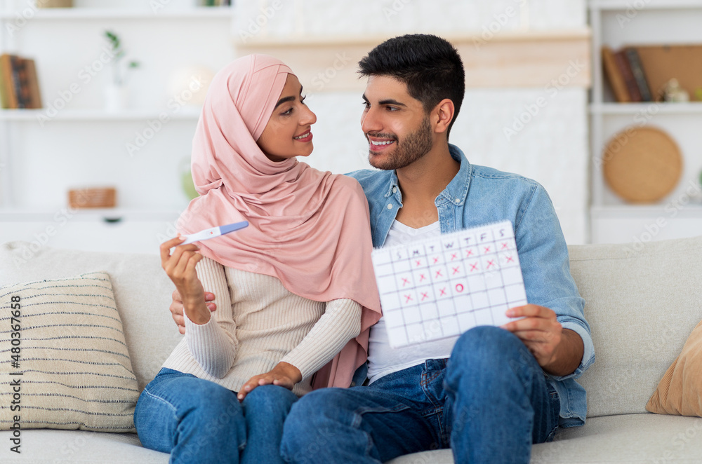 Happy arab couple planning pregnancy, holding positive pregnancy test and ovulation calendar, sitting on sofa