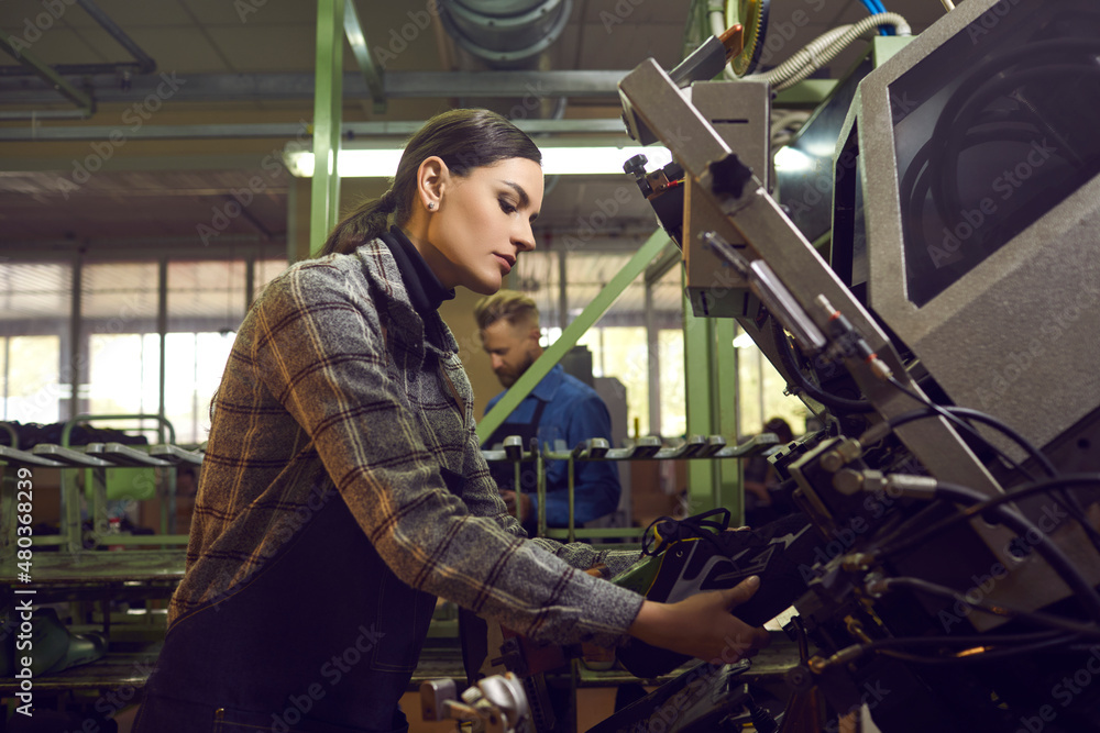 Side view of serious female worker working with special machinery at shoe factory. Woman using industrial press while making new sneakers. Manufacturing industry and mass footwear production concept Side view of serious female worker working with special machinery at shoe factory. Woman using industrial press while making new sneakers. Manufacturing industry and mass footwear production concept