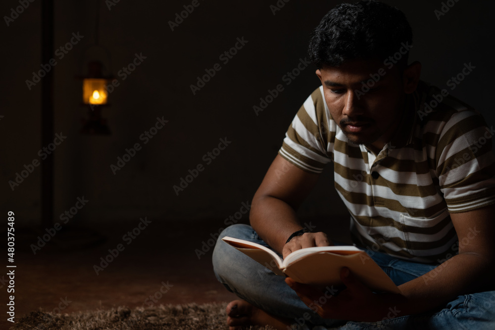 Young student busy reading during night under oil lamp or lantern due to power loss and Poverty - concpet of power cut, blackout during examination.