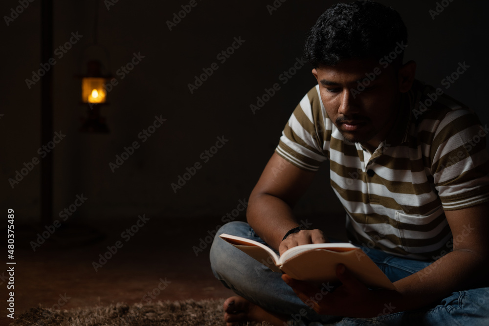 Young student busy reading during night under oil lamp or lantern due to power loss and Poverty - concpet of power cut, blackout during examination. Young student busy reading during night under oil lamp or lantern due to power loss and Poverty - concpet of power cut, blackout during examination.