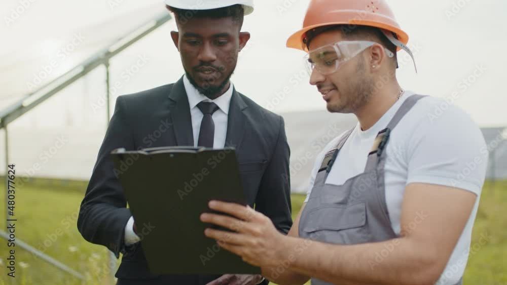 Close up of african man in suit signing documents on clipboard that holding indian engineer. Two multiracial people standing among solar farm and making agreement. Men and technician among solar cells Close up of african man in suit signing documents on clipboard that holding indian engineer. Two multiracial people standing among solar farm and making agreement. Men and technician among solar cells