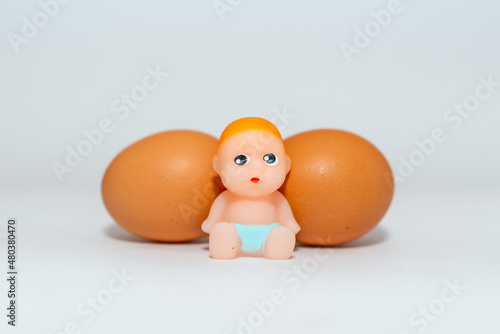 Miniature newborn baby toy and two eggs sitting on a white gray background.Selective focus,copy space.