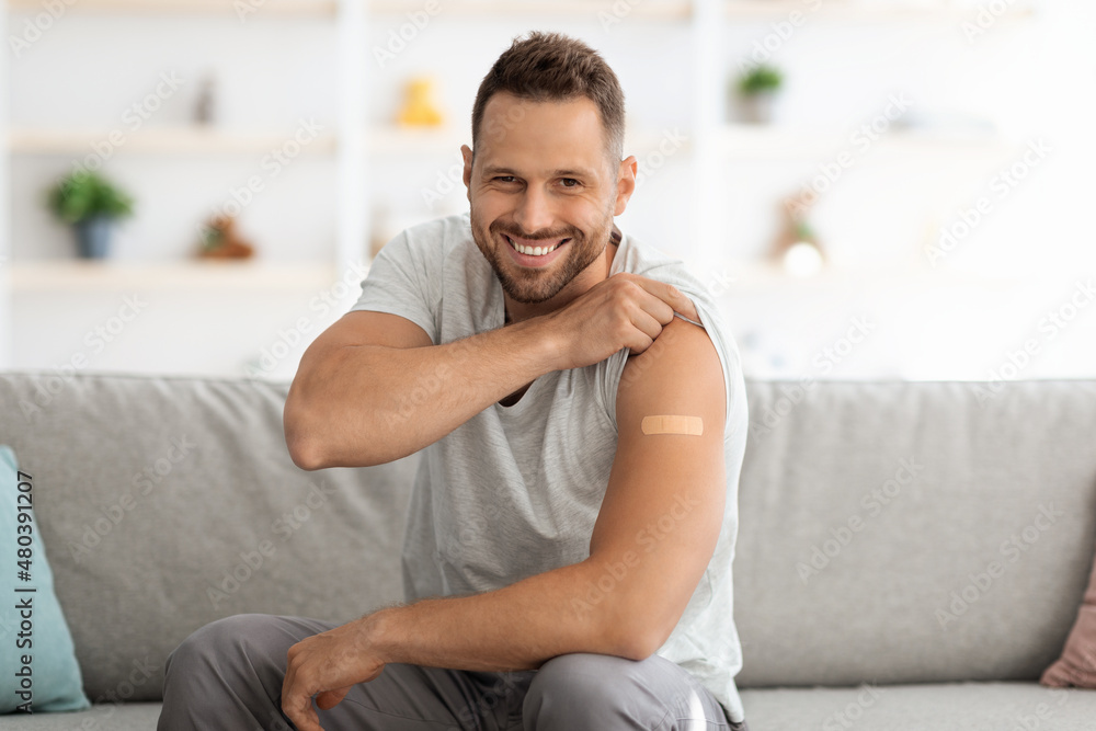 Happy young vaccinated man showing shoulder with medical plaster after vaccine injection and smiling widely to camera