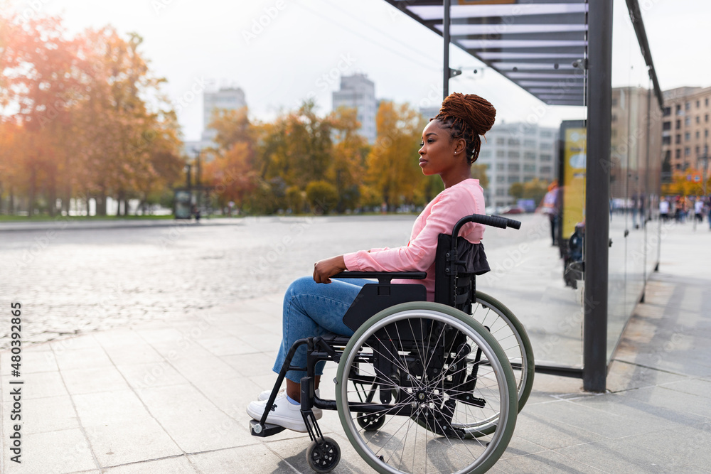 Black paraplegic woman in wheelchair waiting on bus stop, cannot board vehicle suitable for handicapped persons - obrazy, fototapety, plakaty 