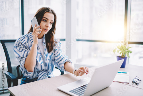 Smart casual female employee using smartphone app for making service consultancy during laptop programming in modern workspace, Caucasian woman connecting to 4g for phoning and call talking via mobile photo