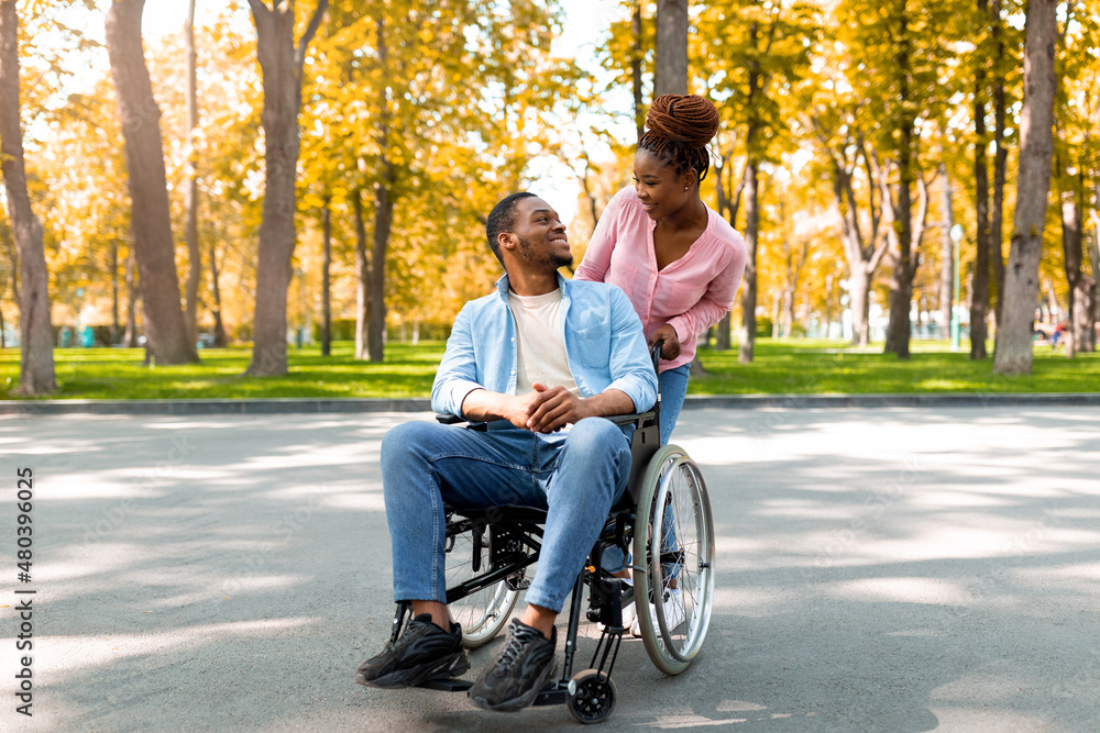 Affectionate black woman and her disabled boyfriend in wheelchair looking at each other on walk at park in autumn Affectionate black woman and her disabled boyfriend in wheelchair looking at each other on walk at park in autumn