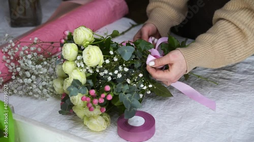 Florist woman creating bouquet at flower shop, small business. Close-up female hands tying a pink ribbon on a fresh bouquet. White roses. Slow motion footage.