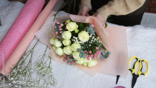 Process of creating bouquet in flower shop, close up. Woman hands working with wrapped paper and ribbon. Florist makes a bouquet of roses. Top view.