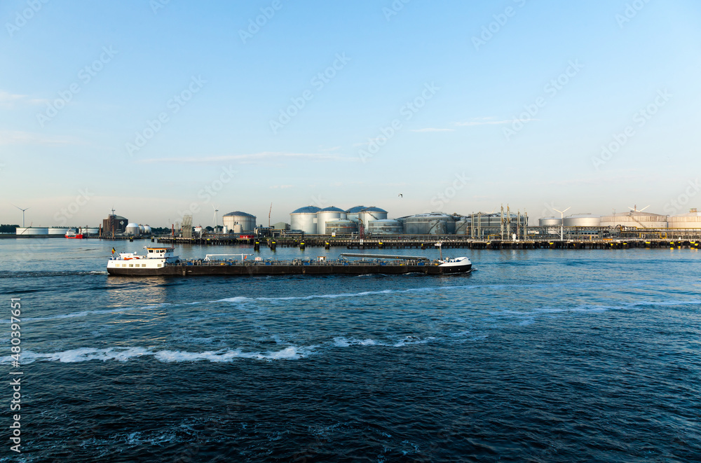 An oil refinery in Amsterdam is near the water and a small refueling tanker passes by.