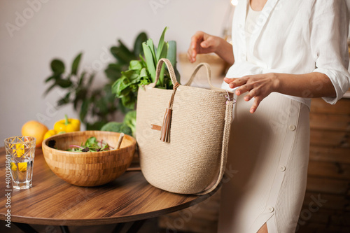 Unrecognizable woman in white shirt taking out green vegetables out of eco reusable bag after grocery shopping for healthy food. 