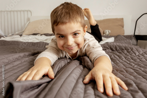 Portrait of little, smiling caucasian boy on the bed at home. Cute child relaxing, resting in bedroom. Positive emotions. Cozy and modern interior. Natural, earth colors. Casual clothes.