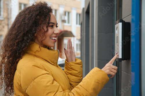 Young African-American woman ringing intercom while waving to camera near building entrance photo