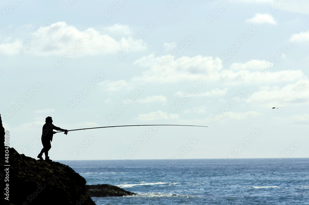 angler fisher in silhouette fishing with rod and reel in atlantic coast with blue sky and clouds , Setubal , Portugal angler fisher in silhouette fishing with rod and reel in atlantic coast with blue sky and clouds , Setubal , Portugal