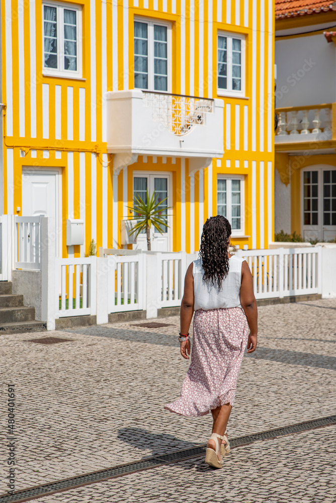 Young African American woman walks the streets with colorful striped houses, Costa Nova, Aveiro, Portugal
