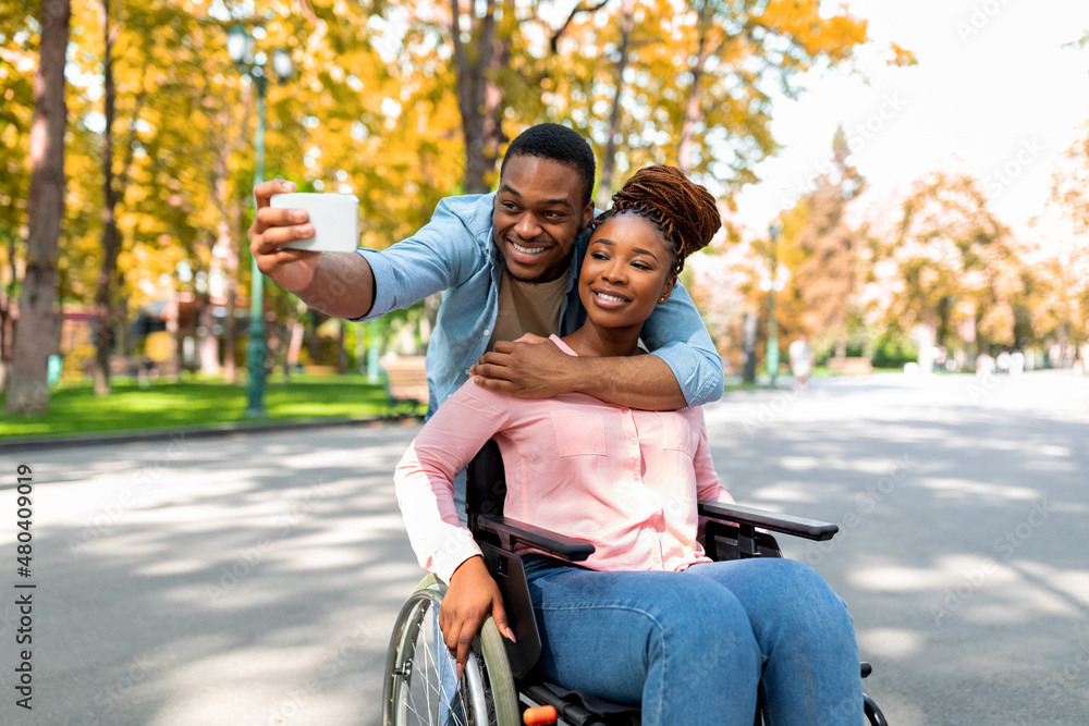 Cheerful black guy hugging his disabled girlfriend in wheelchair, making selfie on smartphone at autumn park Cheerful black guy hugging his disabled girlfriend in wheelchair, making selfie on smartphone at autumn park