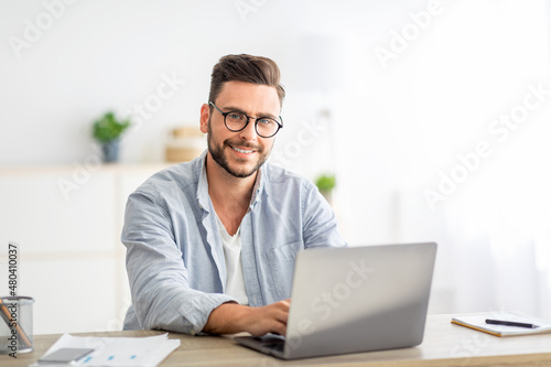 Portrait of freelancer man sitting at desk with laptop computer at home office, smiling at camera, free space