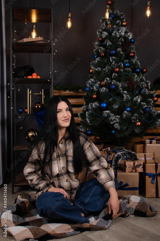 confident beautiful young woman in checkered shirt and jeans near christmas tree decorated in blue tones.