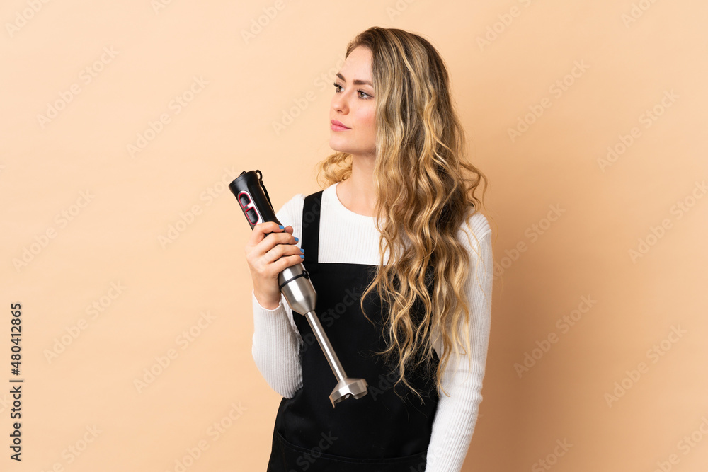Young brazilian woman using hand blender isolated on beige background looking to the side Young brazilian woman using hand blender isolated on beige background looking to the side