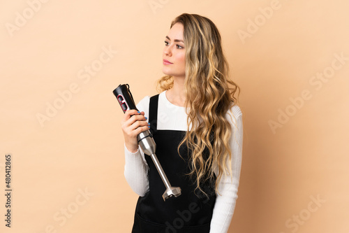 Young brazilian woman using hand blender isolated on beige background looking to the side