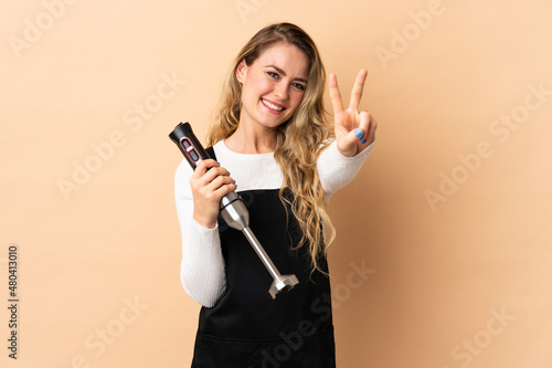 Young brazilian woman using hand blender isolated on beige background smiling and showing victory sign
