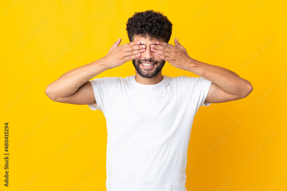 Young Moroccan man isolated on yellow background covering eyes by hands and smiling Young Moroccan man isolated on yellow background covering eyes by hands and smiling