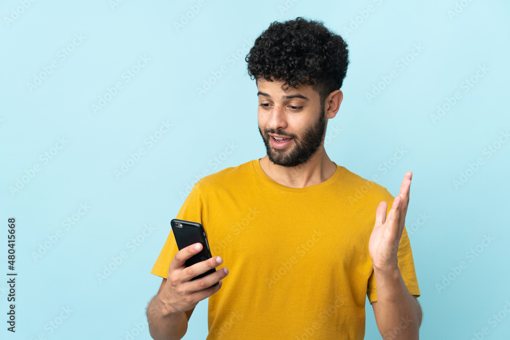 Young Moroccan man isolated on blue background looking at the camera while using the mobile with surprised expression Young Moroccan man isolated on blue background looking at the camera while using the mobile with surprised expression