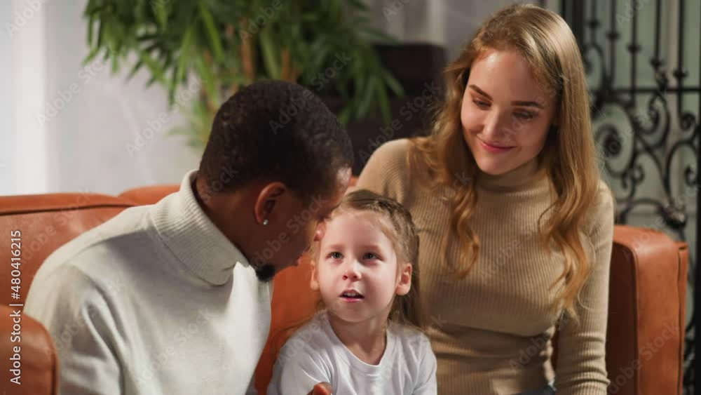 Little daughter talks to African-American father looking at vintage family photo album with blonde long haired mother in sitting room closeup slow motion Little daughter talks to African-American father looking at vintage family photo album with blonde long haired mother in sitting room closeup slow motion