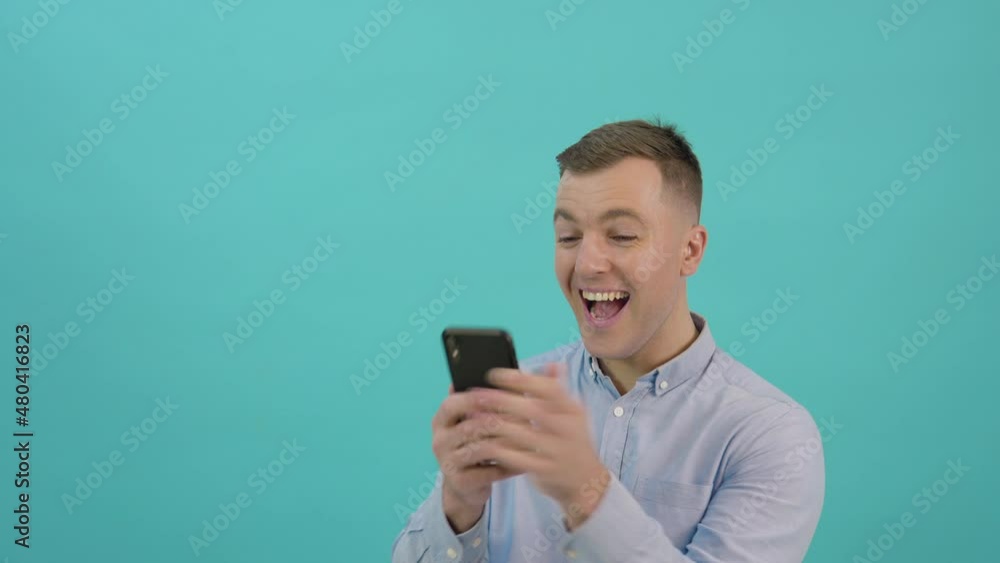 A man in a blue shirt is corresponding on a smartphone and expresses great joy and surprise. Office worker with a mobile phone in his hands stands in front of a bright blue background A man in a blue shirt is corresponding on a smartphone and expresses great joy and surprise. Office worker with a mobile phone in his hands stands in front of a bright blue background