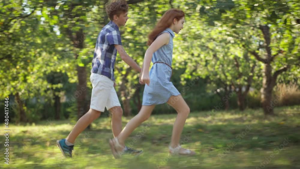 Side view wide shot happy loving teenage couple holding hands running in sunshine outdoors. Tracking shot Caucasian redhead boy and girl having fun dating in spring summer park. First love and joy Side view wide shot happy loving teenage couple holding hands running in sunshine outdoors. Tracking shot Caucasian redhead boy and girl having fun dating in spring summer park. First love and joy
