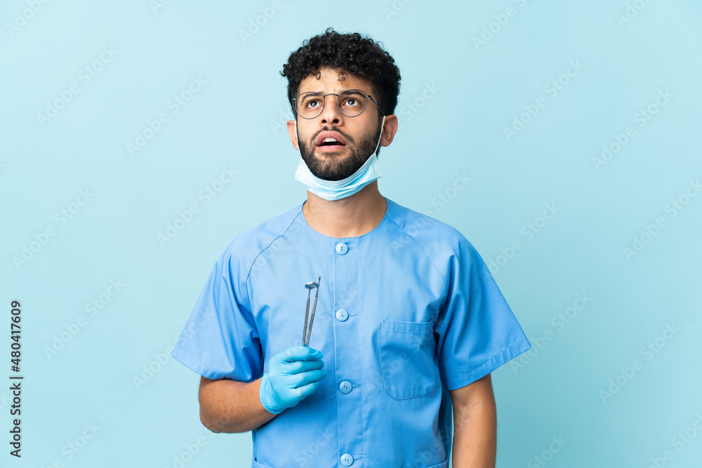 Moroccan dentist man holding tools isolated on blue background looking up and with surprised expression Moroccan dentist man holding tools isolated on blue background looking up and with surprised expression