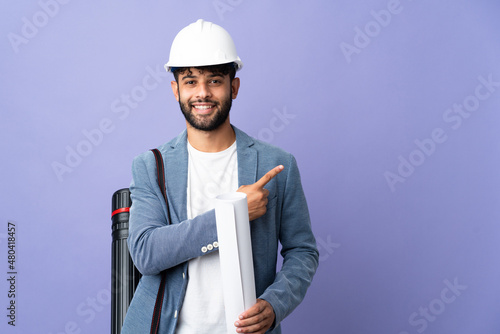Young architect Moroccan man with helmet and holding blueprints over isolated background pointing to the side to present a product