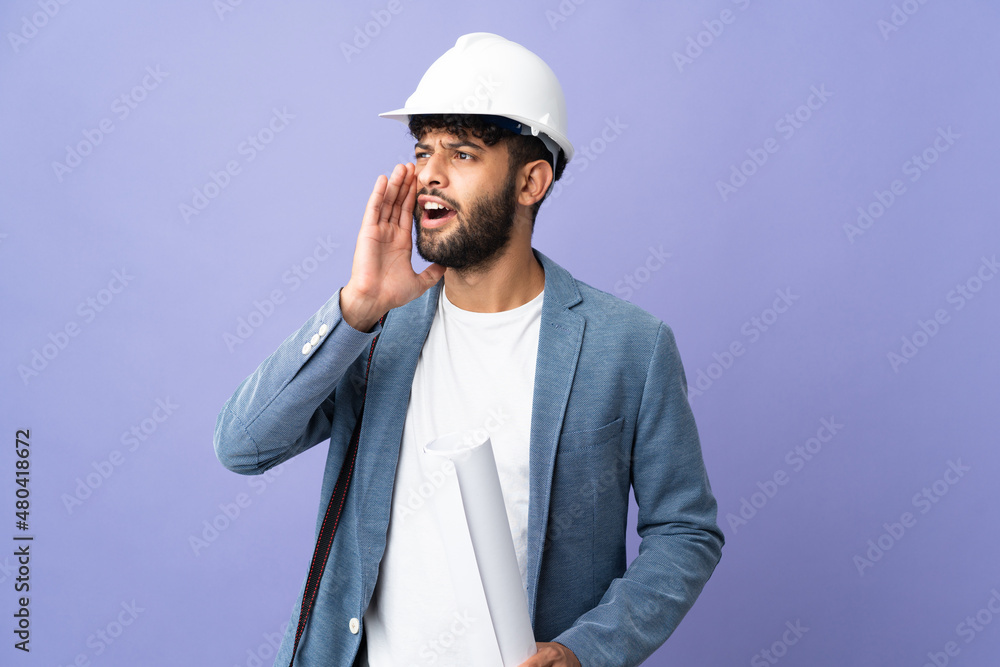 Young architect Moroccan man with helmet and holding blueprints over isolated background shouting with mouth wide open to the side Young architect Moroccan man with helmet and holding blueprints over isolated background shouting with mouth wide open to the side