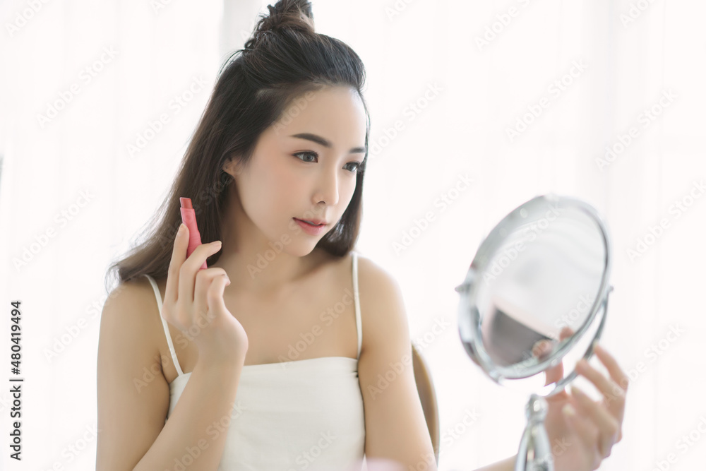 Beautiful young Asian woman applying red lip tint while sitting at table and looking at mirror. Korean girl blogger with excellent makeup with hair tied in knot. Skincare Natural beauty concept.