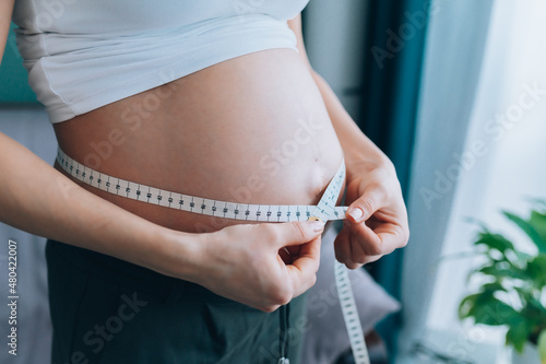 Close-up of torso of unrecognisable pregnant woman in white T-shirt measuring her growing abdomen size with measuring tape at home. Health care pregnancy, weight control concept