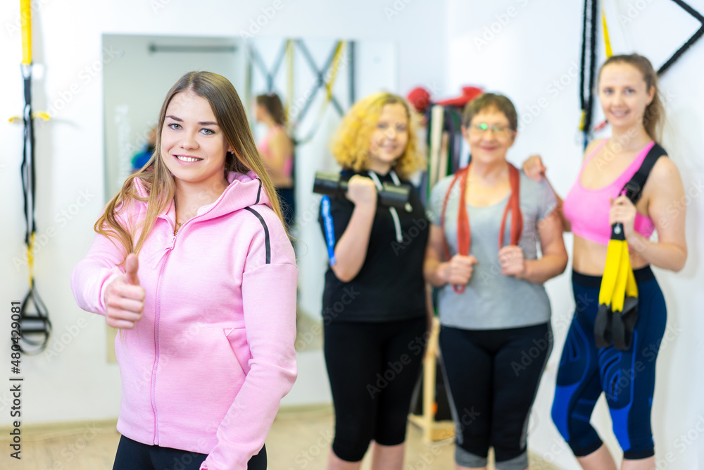 women at the gym. the girl-trainer shows Like and smiles, in the background a girl, grandmother and a woman in a sports uniform are smiling with sports equipment. family. healthy lifestyle