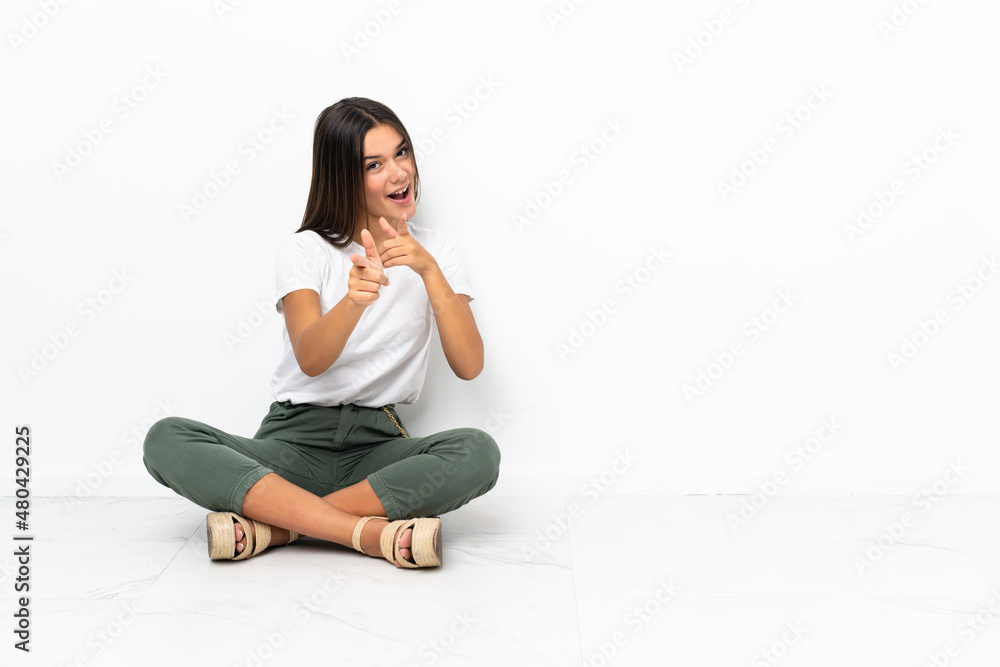Teenager girl sitting on the floor pointing to the front and smiling