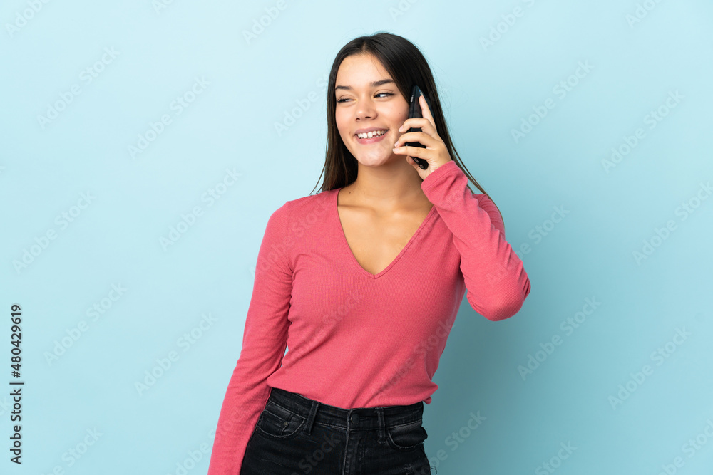 Teenager girl isolated on blue background keeping a conversation with the mobile phone with someone Teenager girl isolated on blue background keeping a conversation with the mobile phone with someone