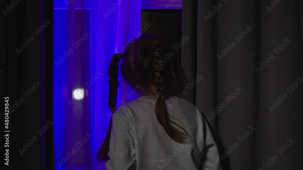 Curious little child with plaits looks out of window attracted by blinking flashlights on street from dark room at night backside view slow motion Curious little child with plaits looks out of window attracted by blinking flashlights on street from dark room at night backside view slow motion