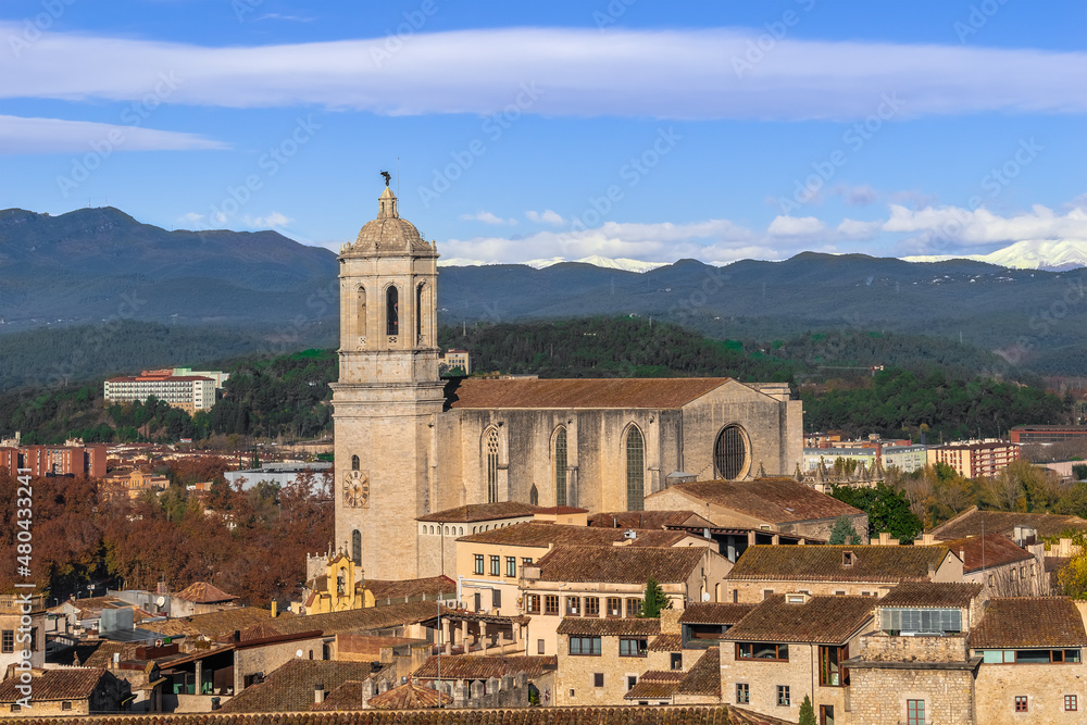 Top view of the old town of Girona with the main building of the Cathedral of Saint Mary against the backdrop of the mountains. The belfry of a Roman Catholic church rises among the brown tiled roofs