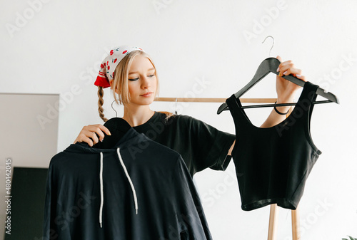 Young lady chossing what to buy, holding hangers with top and hoodie in designer store photo