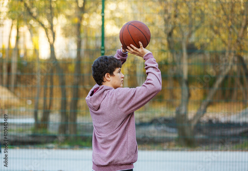 Cute teenager in a hoodie playing basketball. Young boy with ball learning dribble and shooting on the city court. Hobby for kids, active lifestyle © Natali