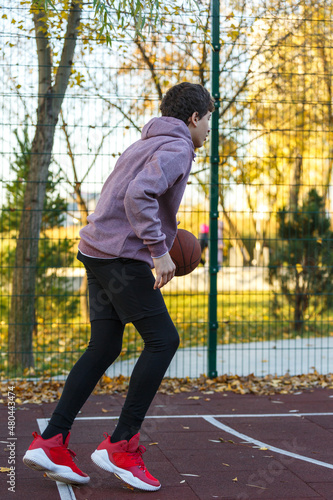 Cute teenager in a hoodie playing basketball. Young boy with ball learning dribble and shooting on the city court. Hobby for kids, active lifestyle
