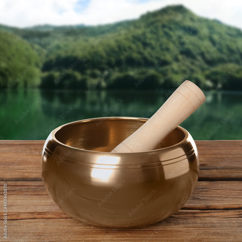 Golden singing bowl and mallet on wooden table against mountain landscape Golden singing bowl and mallet on wooden table against mountain landscape