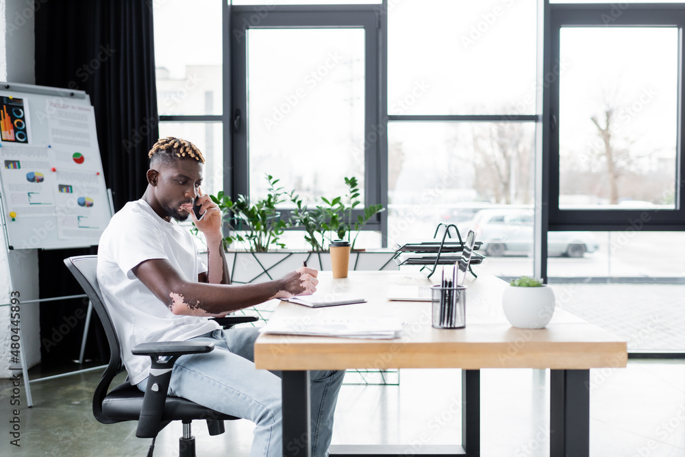 african american man with vitiligo talking on mobile phone near laptop and flip chart in office. african american man with vitiligo talking on mobile phone near laptop and flip chart in office.
