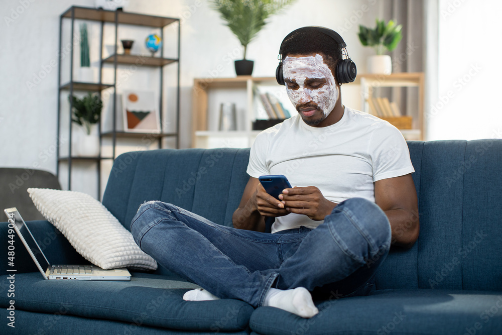 Young handsome African American man with facial mask on face, wearing headphones, sitting on sofa at home and listening to music choosing favourite song from smartphone playlist